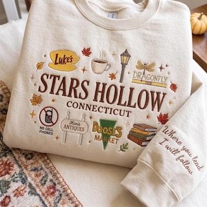 May include: A cream-colored crewneck sweatshirt featuring brown embroidered text that reads Stars Hollow Connecticut. The design includes icons of a coffee cup, a street lamp, a dragonfly, books, and signs for Luke's, Kim's Antiques, and Doose's Market.