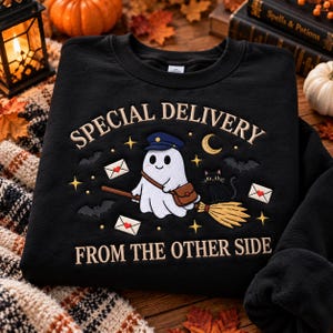 May include: A black crewneck sweatshirt featuring an embroidered design of a white ghost wearing a blue mail carrier hat and a brown satchel, riding a broomstick. The text reads SPECIAL DELIVERY FROM THE OTHER SIDE, with a black cat, bats, and envelopes.