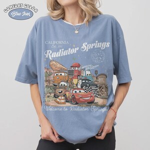 May include: Blue t-shirt with a graphic featuring characters from the movie Cars, including Lightning McQueen. The shirt has the text "California Est. 2012 Radiator Springs" and "Welcome to Radiator Springs". The shirt is labeled "Comfort Color Blue Jean".
