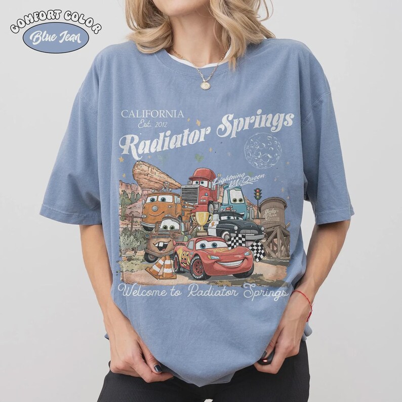 May include: Blue t-shirt with a graphic featuring characters from the movie Cars, including Lightning McQueen. The shirt has the text "California Est. 2012 Radiator Springs" and "Welcome to Radiator Springs". The shirt is labeled "Comfort Color Blue Jean".