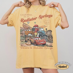 May include: Mustard yellow t-shirt featuring a graphic of characters from the movie Cars, with the text "Radiator Springs" and "Welcome to Radiator Springs". The shirt also has the text "California Est. 2012".