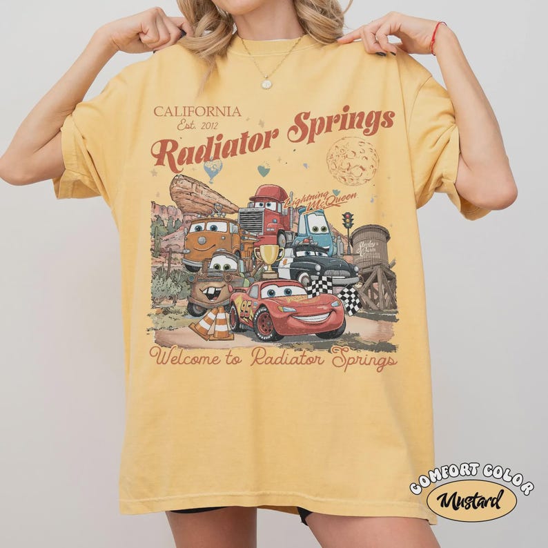 May include: Mustard yellow t-shirt featuring a graphic of characters from the movie Cars, with the text "Radiator Springs" and "Welcome to Radiator Springs". The shirt also has the text "California Est. 2012".