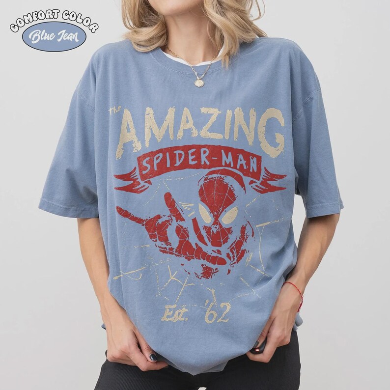 May include: Blue jean Comfort Colors t-shirt featuring a distressed graphic of Spider-Man with the text "The Amazing Spider-Man" and "Est. '62". The graphic is in red and tan colors. The shirt has short sleeves and a relaxed fit.