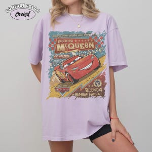 May include: A light purple t-shirt featuring a graphic of Lightning McQueen from the Disney Pixar Cars movie. The design includes the text "Lightning McQueen" and "Radiator Springs Saturday Night Special". The shirt has short sleeves and a relaxed fit.