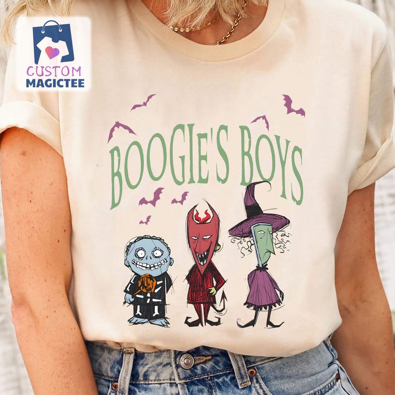 May include: Beige t-shirt featuring the text "BOOGIE'S BOYS" in green and purple, with cartoon illustrations of three characters and bats. The characters include a blue zombie, a red devil, and a green witch. The shirt has short sleeves.