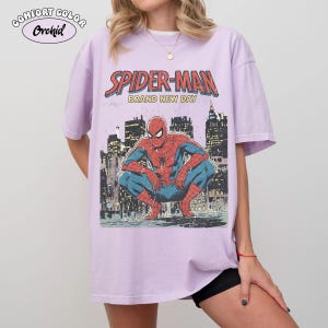 May include: A light purple t-shirt featuring a graphic of Spider-Man in a crouching pose against a cityscape backdrop. The text "SPIDER-MAN BRAND NEW DAY" is printed above the image. The shirt has short sleeves and a relaxed fit.
