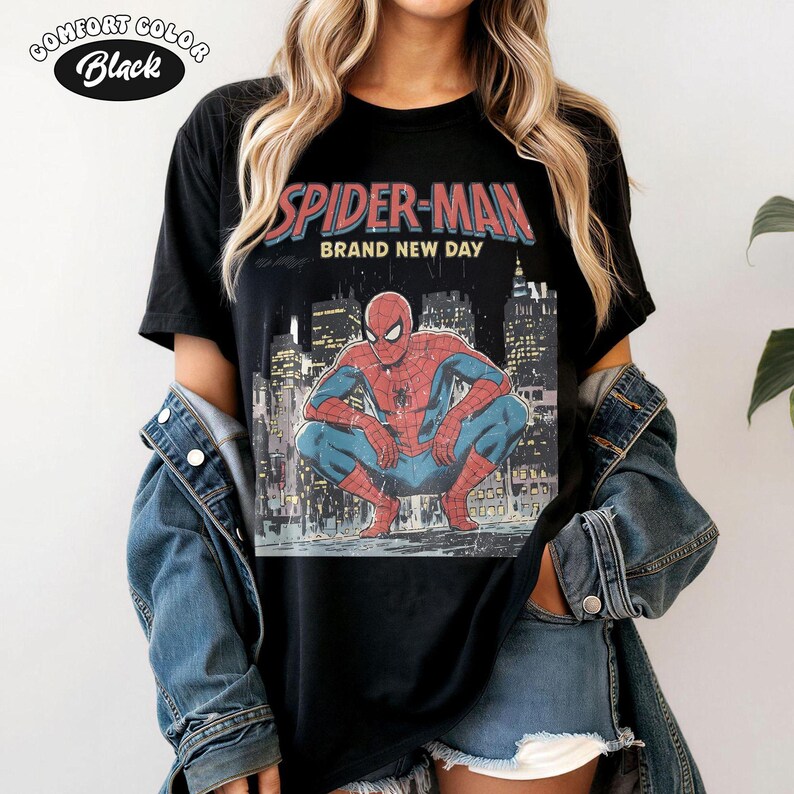 May include: Black t-shirt featuring a graphic of Spider-Man in a crouching pose against a cityscape backdrop. The text "SPIDER-MAN BRAND NEW DAY" is printed above the image in red and blue. The shirt is labeled "Comfort Color Black".