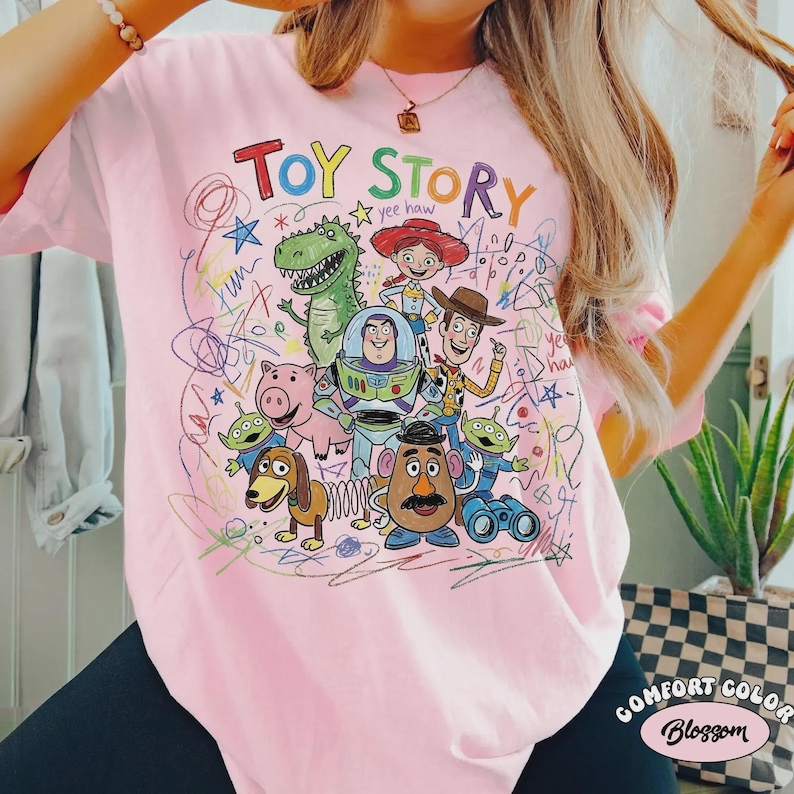 May include: Pink t-shirt featuring a colorful graphic of Toy Story characters, including Woody, Buzz Lightyear, and Rex. The words "Toy Story" are written in a playful font above the characters. The shirt is a Comfort Colors brand.