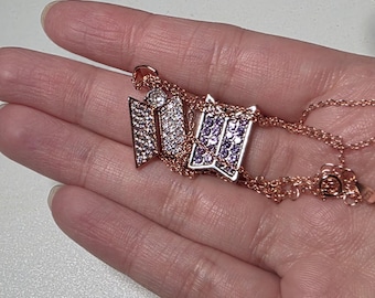 May include: Rose gold necklace featuring a pendant with three stylized, open book designs. The pendants are embellished with clear and purple crystals. The necklace is displayed on a hand.