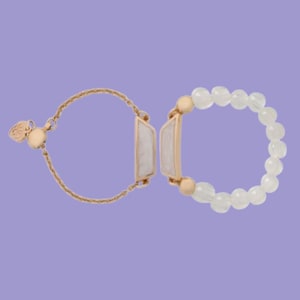 May include: Two rings on a lavender background. One ring is a beaded design with white beads. The other ring has a gold chain and a rectangular white stone with gold trim. Jewelry.
