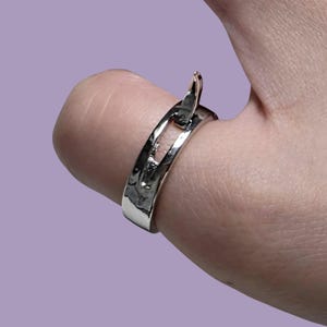May include: A polished silver-toned ring featuring an open, split-band design with a curved, pointed element extending from the top. The metal surface has a smooth, reflective finish that catches the light, creating a sleek and continuous appearance.