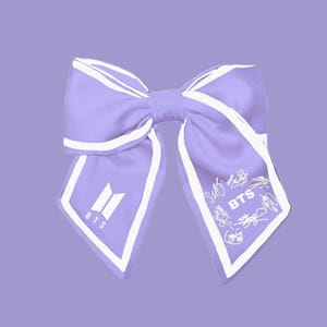 May include: A lilac satin bow with white edging, featuring the BTS logo and signatures. The bow is a light purple colour and has a classic bow shape. The BTS logo is in white.