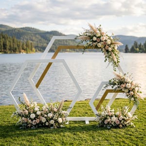 Può includere: Un arco nuziale geometrico bianco e oro decorato con composizioni floreali. L'arco è posizionato su un'area erbosa con un lago e montagne sullo sfondo. Le composizioni floreali includono rose bianche e rosa, erba della pampa e verde.