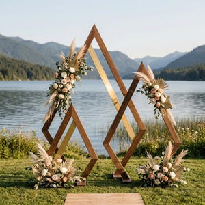May include: A wooden wedding arch with a geometric design, featuring diamond and triangle shapes. The arch is decorated with floral arrangements in shades of white, cream, and peach, with accents of pampas grass. The background includes a lake and mountains.