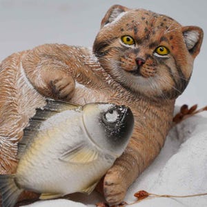 Op de afbeelding: Gedetailleerd beeldje van een Pallas' kat, met bruine en bruingele vacht, die een vis vasthoudt. De kat heeft gele ogen en ligt in een besneeuwde omgeving. De vis is zilver en wit.