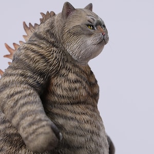 Puede incluir: Figura detallada de una criatura felina con un diseño inspirado en Godzilla. La figura es principalmente gris con rayas más oscuras y tiene una aleta dorsal espinosa. El gato tiene ojos amarillos y una expresión decidida.