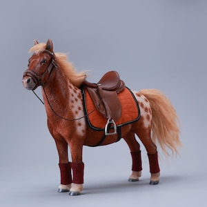 Op de afbeelding: Gedetailleerd model van een paard met een bruin-witte gevlekte vacht, compleet met een bruin zadel, hoofdstel en rode beenbeschermers. Het paard heeft een vloeiende blonde manen en staart, tegen een neutrale grijze achtergrond.