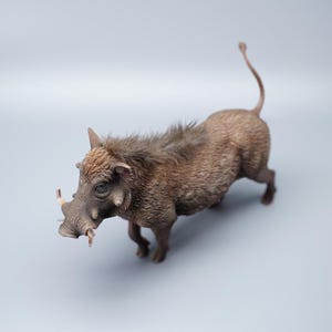 May include: A detailed figurine of a warthog, featuring a textured, brown body with visible bristles and tusks. The animal is posed in a walking stance, with its tail curled upwards. The figurine is set against a plain, light gray background.