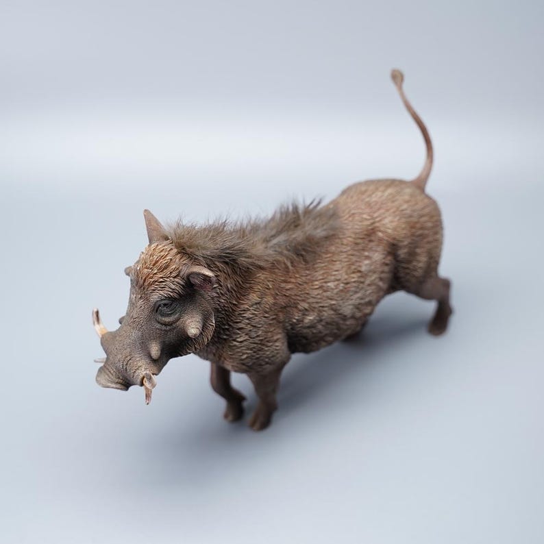 May include: A detailed figurine of a warthog, featuring a textured, brown body with visible bristles and tusks. The animal is posed in a walking stance, with its tail curled upwards. The figurine is set against a plain, light gray background.