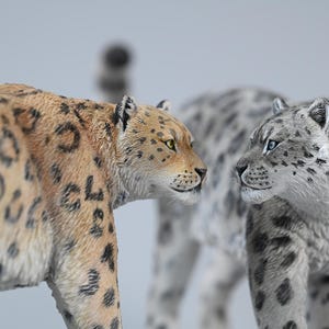 Op de afbeelding: Twee gedetailleerde luipaardbeeldjes. Eén is bruin met zwarte vlekken en gele ogen, de andere is grijs met donkere vlekken en blauwe ogen. De beeldjes kijken elkaar aan, met een derde, wazige luipaard op de achtergrond.