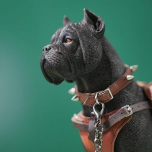 Pode incluir: Estatueta de cão preto com coleira e arnês de couro castanho. A coleira apresenta pontas prateadas. O cão tem olhos cor de laranja, sobre um fundo verde.