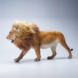 Op de afbeelding: Een levensechte leeuwenfiguur met een bruin lichaam en een lichtbruine manen. De leeuw wordt afgebeeld in een wandelhouding, met zijn mond lichtjes open. De figuur staat tegen een effen, lichte achtergrond.