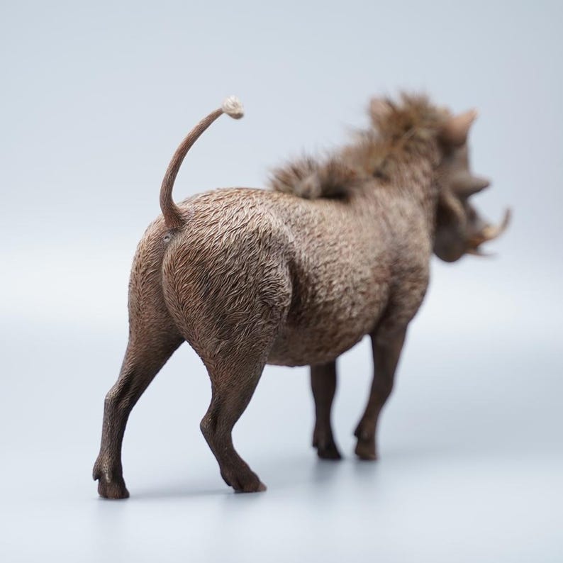 May include: A detailed figurine of a warthog, viewed from the rear. The sculpture is primarily brown with textured fur and visible tusks. The tail is curved upwards, and the legs are positioned as if the animal is standing.