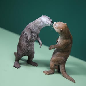 Op de afbeelding: Twee otterbeeldjes, één grijs en één bruin, in een aandoenlijke pose. Ze staan rechtop, met hun neuzen bijna tegen elkaar. De achtergrond is een verloop van groen, wat een naturalistische achtergrond creëert voor deze dierensculpturen.