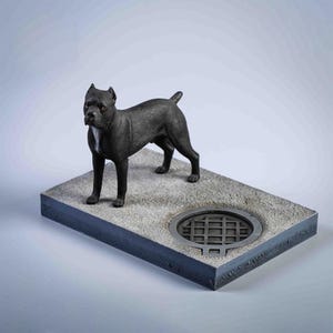 May include: A black dog statue stands on a rectangular, grey concrete base with a manhole cover detail. The dog has a short coat. The base has dark grey edges. The statue is a decorative item.