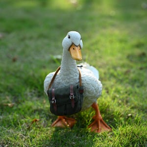 Op de afbeelding: Een witte eendenfiguur met een gele snavel en oranje voeten, met een kleine groene canvas tas met bruine riemen. De eend staat op groen gras.