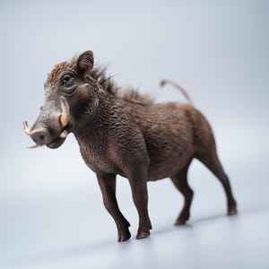 May include: A detailed figurine of a warthog, showcasing a brown, textured coat and prominent tusks. The sculpture captures the animal's distinctive features, including its bristly mane and curved tail, set against a soft, neutral background.