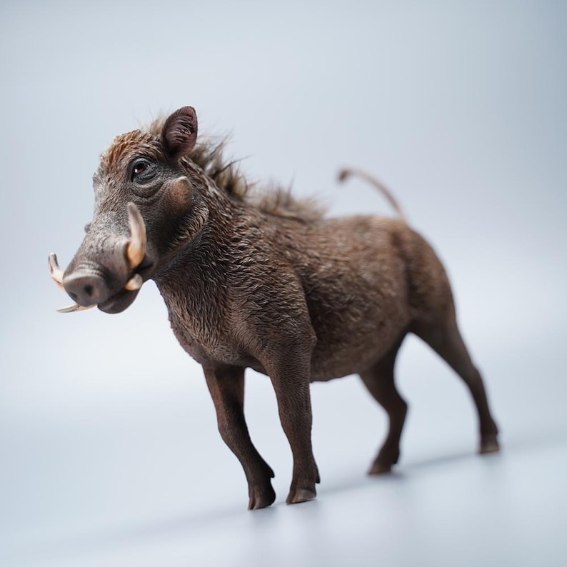May include: A detailed figurine of a warthog, showcasing a brown, textured coat and prominent tusks. The sculpture captures the animal's distinctive features, including its bristly mane and curved tail, set against a soft, neutral background.