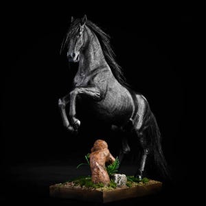 Op de afbeelding: Een zwart-wit sculptuur van een steigerend paard met vloeiende manen en staart. Het paard staat boven een klein, gedetailleerd diorama met een figuur, groen en een rots. De achtergrond is effen zwart.