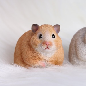 Op de afbeelding: Een gedetailleerd, oranjebruin hamsterbeeldje met zwarte ogen, een roze neus en zichtbare snorharen. De hamster rust op een witte, zachte ondergrond. Het beeldje heeft een realistisch uiterlijk.