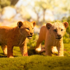 Op de afbeelding: Twee leeuwenwelpen staan in een grasveld. De welpen hebben lichtbruine vacht en kijken de kijker aan. De achtergrond is wazig, met hints van bomen en zonlicht. De welpen lijken zich in een natuurlijke habitat te bevinden.