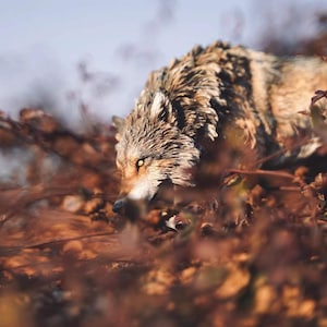 Op de afbeelding: Een gedetailleerde sculptuur van een wolf, met de nadruk op zijn kop en bovenlichaam. De wolf heeft een mix van bruine en grijze vacht, met doordringende gele ogen. Het is omgeven door bruine bladeren, wat een naturalistische scène creëert.
