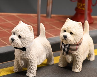 May include: Two cream-colored West Highland White Terrier figurines. One wears a black bow tie, the other a plaid scarf. They stand on a gray surface with a yellow line, suggesting a street scene. A red fire hydrant is visible in the background.