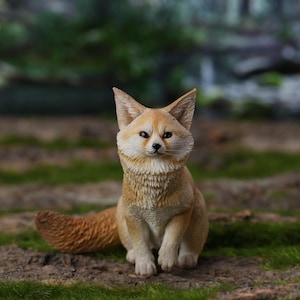 Op de afbeelding: Gedetailleerd beeldje van een fennek, met grote oren en een pluizige staart. De vos is afgebeeld in een zittende positie met een lichtbruin en crèmekleurig kleurenpalet. Het beeldje staat tegen een natuurlijke buitenachtergrond met groen mos.