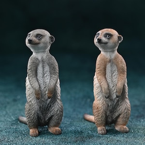 Op de afbeelding: Twee stokstaartjes beeldjes. Eén is grijs, de andere is bruin. Beide staan rechtop met hun armen gekruist. Ze hebben gedetailleerde vachttexturen en staan tegen een turquoise achtergrond.