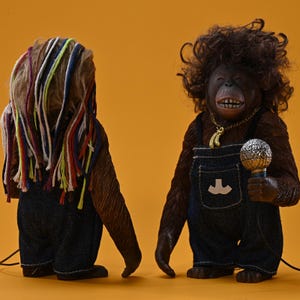 Op de afbeelding: Twee bruine orang-oetan beeldjes. Eén heeft kleurrijk garen haar en kijkt weg. De andere, naar voren gericht, draagt een spijkerbroek met bretels, een gouden ketting met een bananen hanger en houdt een microfoon vast. De achtergrond is oranje.