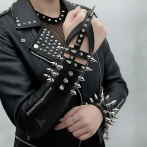 May include: A black leather jacket featuring silver-toned metal studs and a matching black leather hand harness with silver spikes. The jacket has a notched lapel with studs, a zippered sleeve, and a mesh neckline detail, paired with a spiked wrist cuff.
