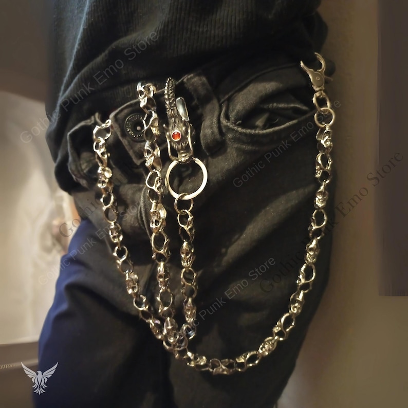 May include: Silver-toned skull chain wallet accessory. The chain features skull links and a dragon-shaped clasp with a red gem. Attached to a pair of black jeans, the chain adds a gothic punk aesthetic.
