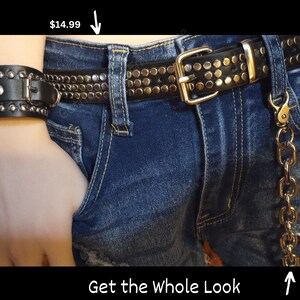 K&ouml;nnte beinhalten: Ein schwarzer Lederg&uuml;rtel mit Nieten, einer silbernen Schnalle und einem passenden schwarzen Lederarmband. Der G&uuml;rtel wird mit einer blauen Jeans und einer goldenen Kette getragen.
