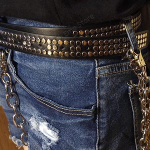 K&ouml;nnte beinhalten: Ein schwarzer Nieteng&uuml;rtel mit einer silbernen Kette daran. Der G&uuml;rtel wird &uuml;ber einer blauen Jeans-Shorts getragen.