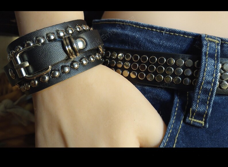 K&ouml;nnte beinhalten: Ein schwarzes Lederarmband mit silbernen Nieten und einer Schnalle. Das Armband wird am Handgelenk einer Person getragen. Die Person tr&auml;gt auch einen schwarzen G&uuml;rtel mit silbernen Nieten.