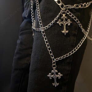 May include: Silver-colored chain belt with two decorative cross pendants. The belt is made of multiple linked chains and hangs over black fabric. The crosses have ornate, gothic-style designs.