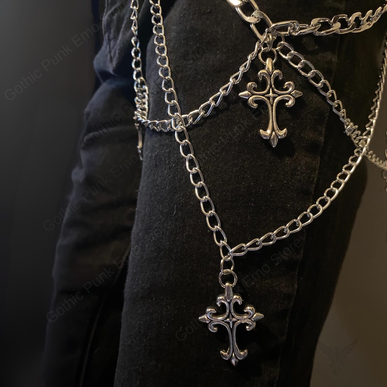 May include: Silver-colored chain belt with two decorative cross pendants. The belt is made of multiple linked chains and hangs over black fabric. The crosses have ornate, gothic-style designs.
