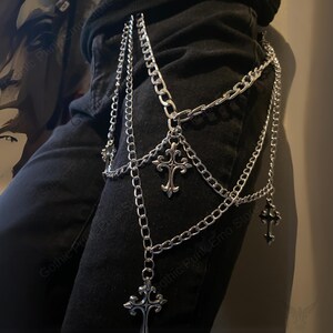 May include: Silver-colored chain belt with multiple strands and decorative cross pendants. The belt is designed to be worn around the waist and upper thigh, adding a gothic or punk aesthetic to an outfit.