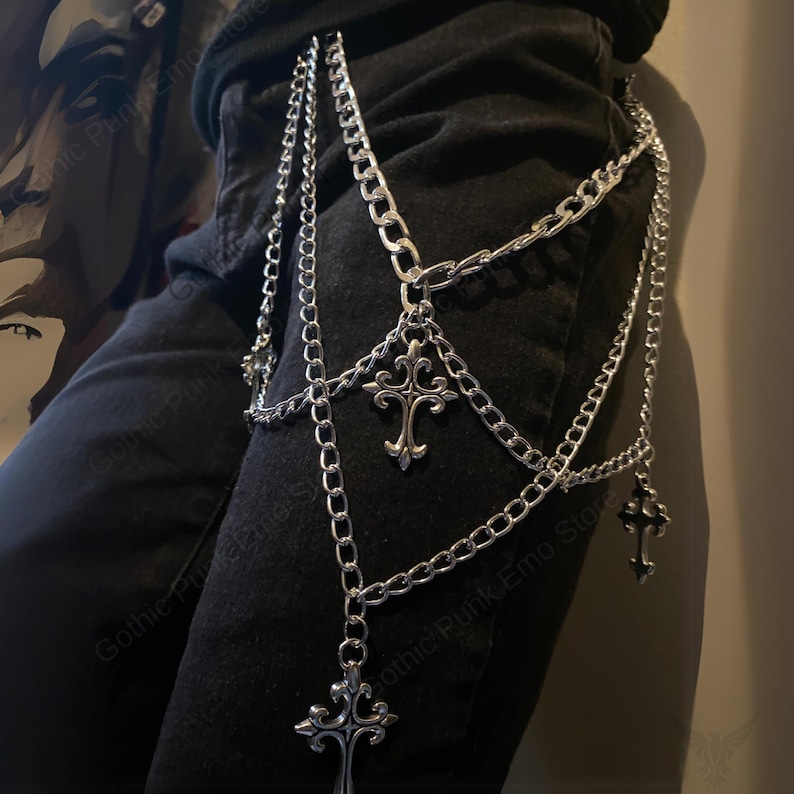 May include: Silver-colored chain belt with multiple strands and decorative cross pendants. The belt is designed to be worn around the waist and upper thigh, adding a gothic or punk aesthetic to an outfit.