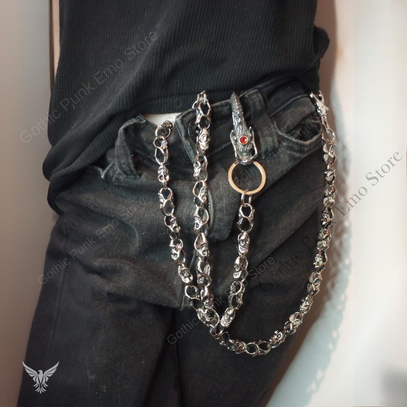 May include: A silver-toned wallet chain with skull and dragon head details. The chain is attached to a belt loop with a dragon head clasp featuring a red gem. The chain is made of multiple skull links.
